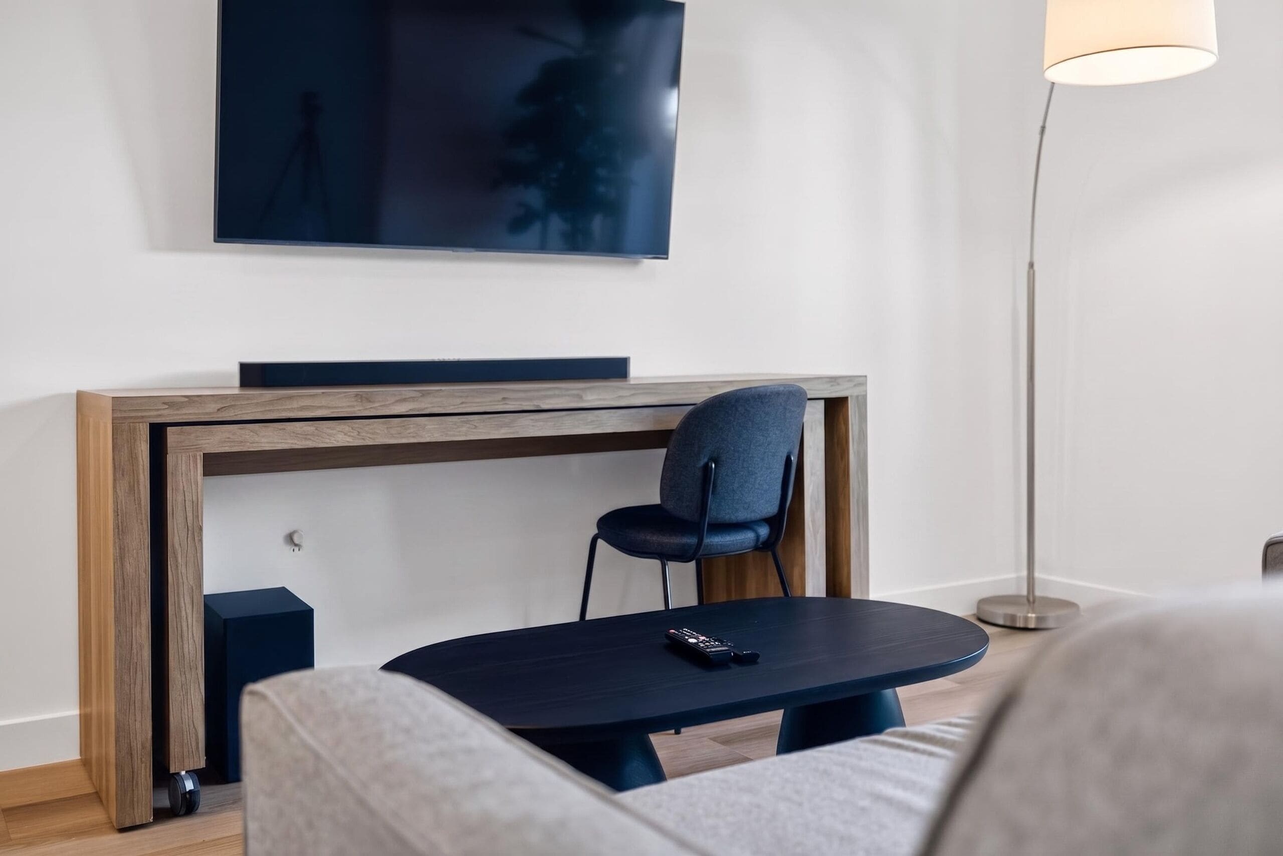 Una habitación moderna con un escritorio de madera, una silla, una mesa redonda, un televisor montado en la pared y una lámpara de pie.