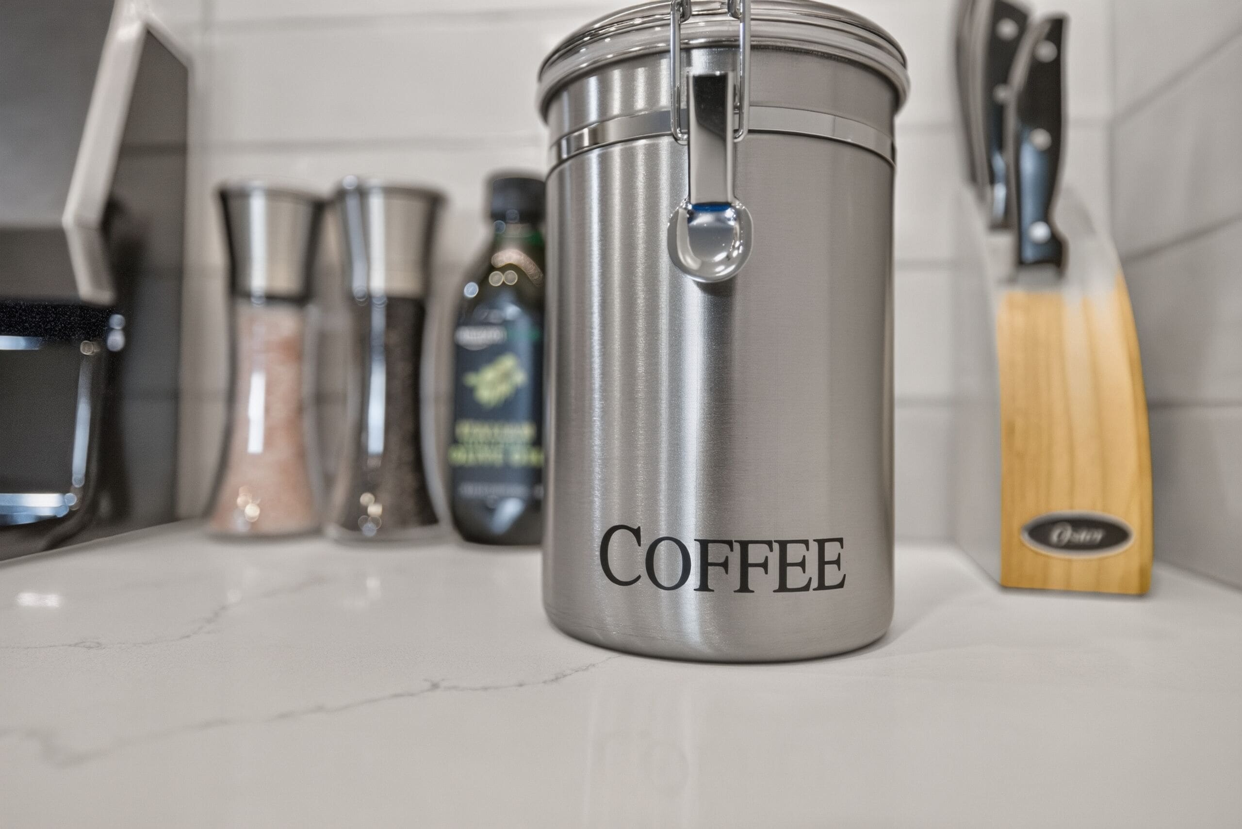 Un récipient à café en acier inoxydable, doté d’une anse, posé sur un plan de travail de cuisine, et sur lequel est inscrit le mot « CAFÉ ».