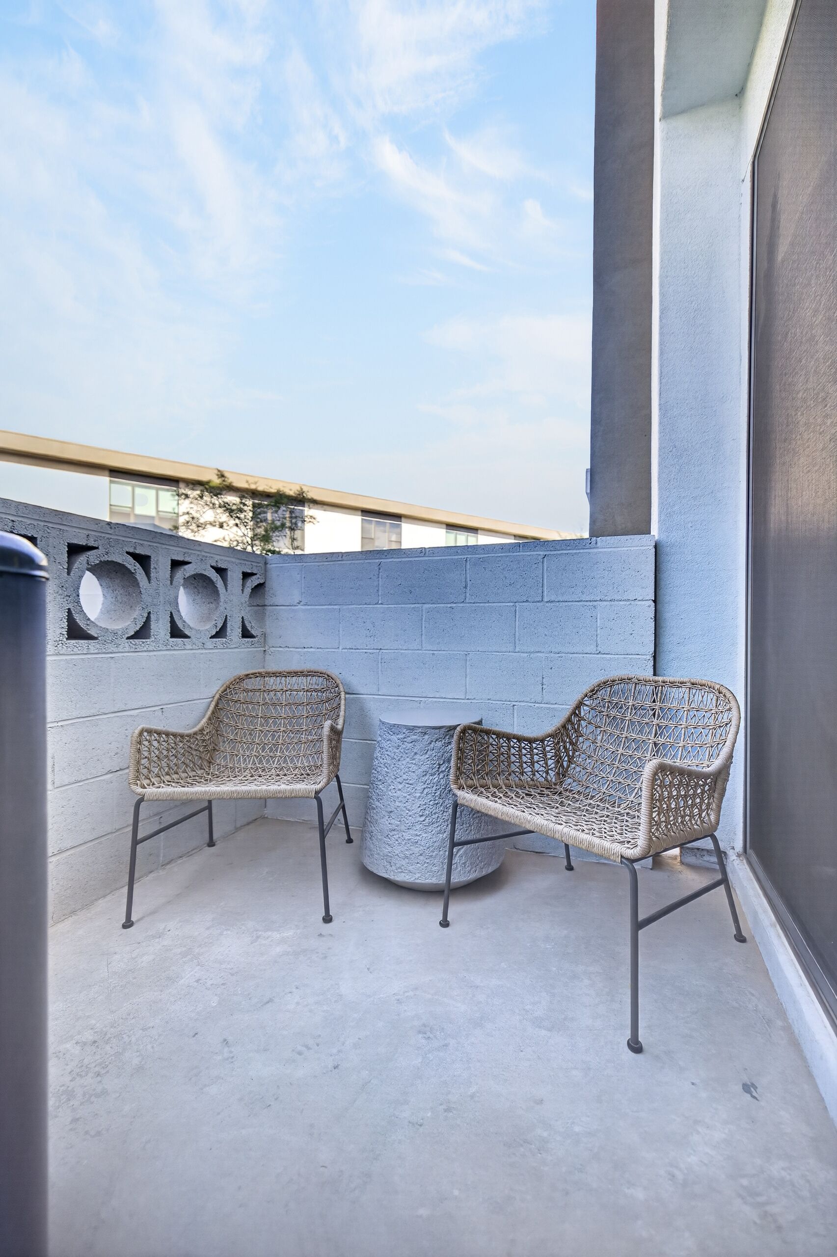 A balcony with two wicker chairs and a small round table, a concrete floor, and a grey wall with a circular design.
