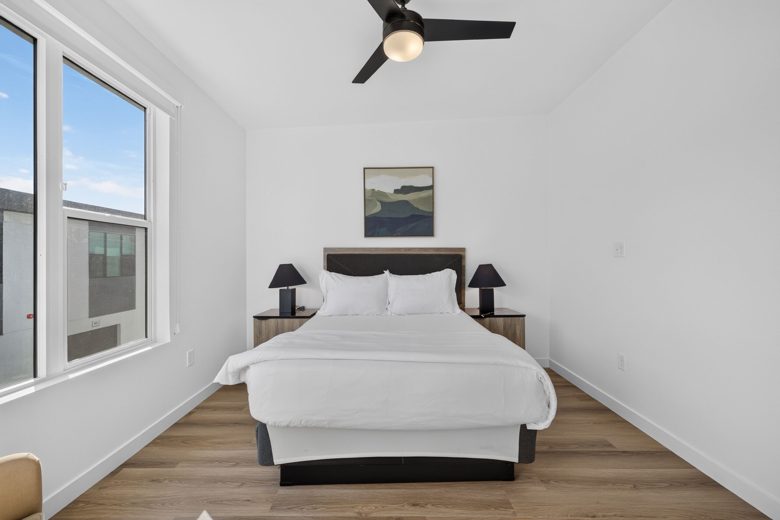 Un dormitorio con cama, ventilador de techo, mesita de noche con lámpara, un cuadro en la pared y una ventana.