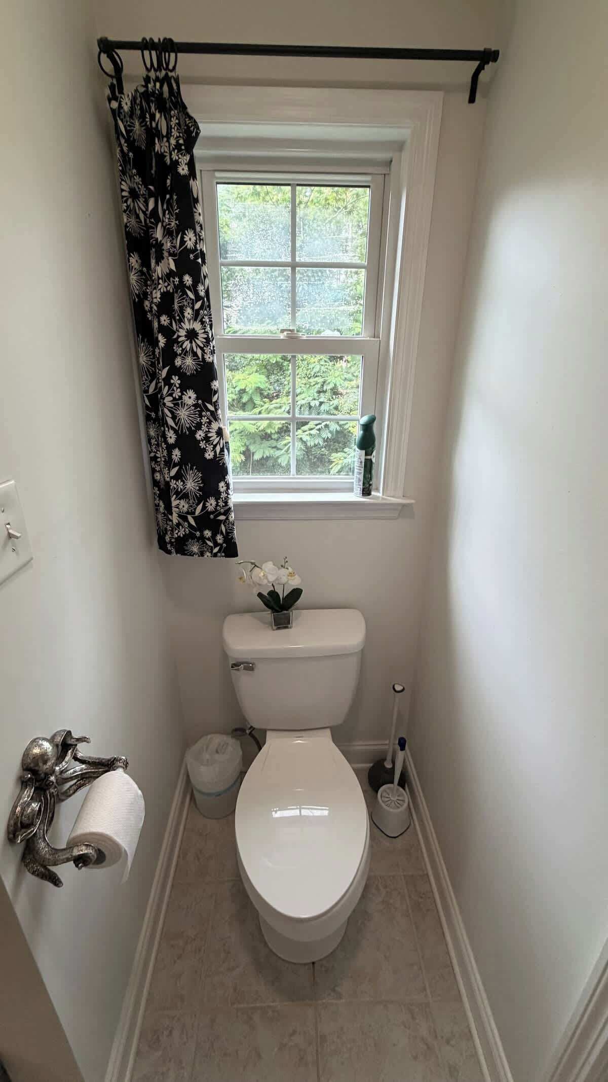 A small bathroom with a toilet, a window with a view of greenery, a shower curtain, and a toilet paper holder.