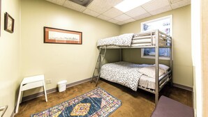 Une chambre avec des lits superposés, un petit bureau, une poubelle, un tapis à motifs et un tableau encadré au mur.