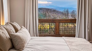 Un dormitorio con una cama grande, ropa de cama color beige y vistas a un paisaje montañoso a través de una ventana con barandilla de madera.