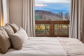 Ein Schlafzimmer mit einem großen Bett, beiger Bettwäsche und Blick auf eine Berglandschaft durch ein Fenster mit Holzgeländer.