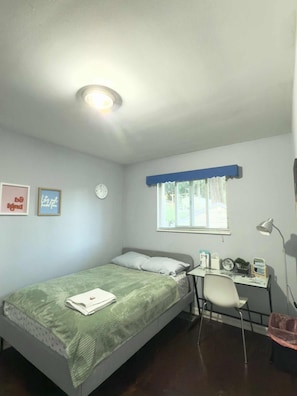 Una cama individual con una manta verde, un escritorio con una silla, una ventana con persianas y un reloj de pared.