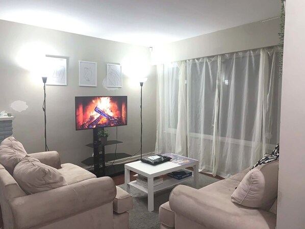 Una sala de estar con televisor de pantalla plana, dos sofás color beige, una mesa de centro y cortinas ligeras.