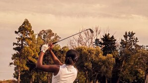 Eine Person spielt Golf auf einem Platz, im Hintergrund sind Bäume zu sehen.