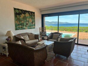 A living room with wicker furniture, a painting on the wall, and a view of the ocean. - Beachfront Home (Bormes-les-Mimosas)
