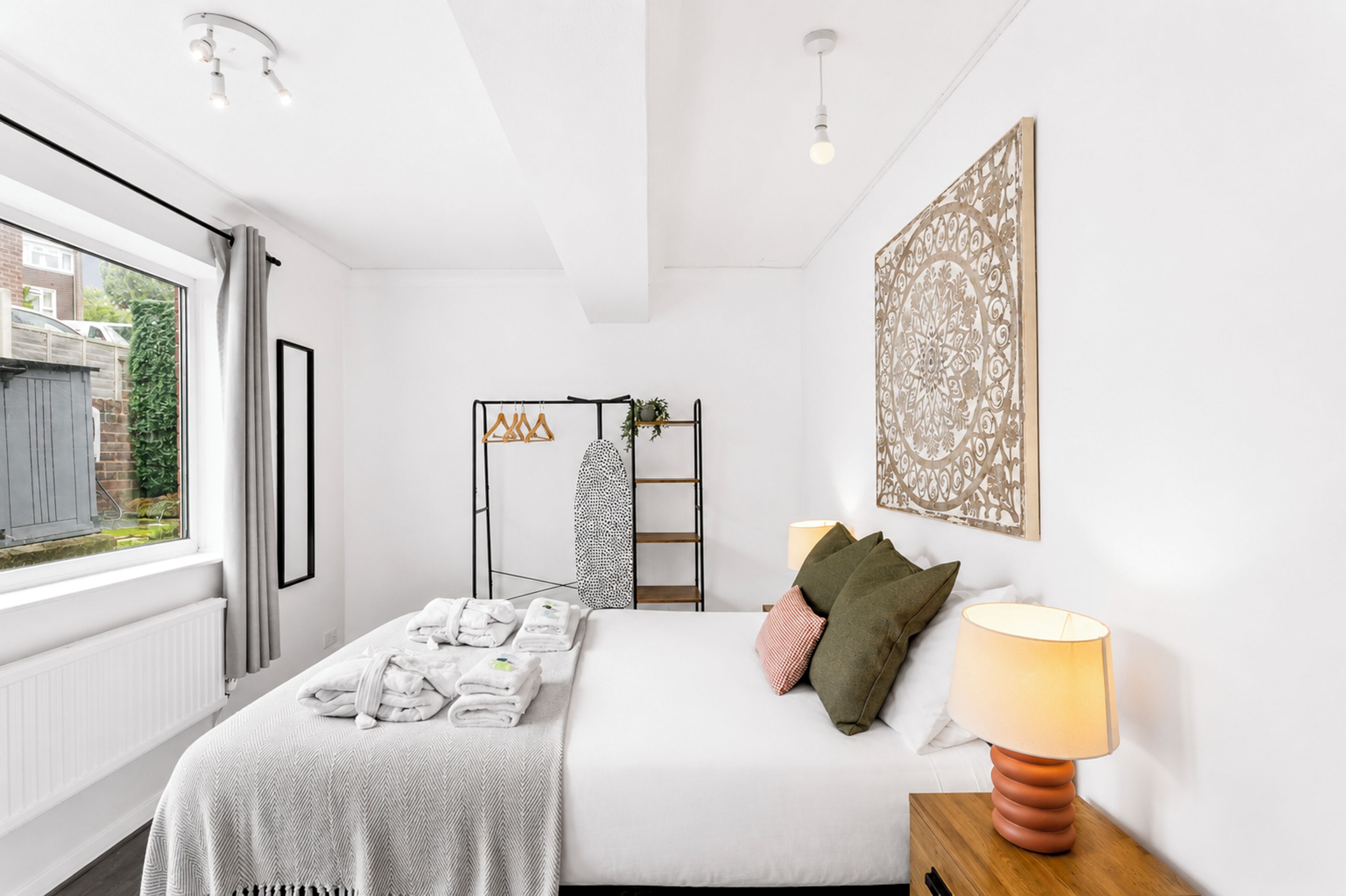 A bedroom with a bed, a bedside lamp, a window with curtains, a radiator, a wall-mounted shelf with towels, and a decorative wall piece.