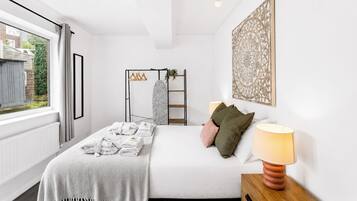 A bedroom with a bed, a bedside lamp, a window with curtains, a radiator, a wall-mounted shelf with towels, and a decorative wall piece.
