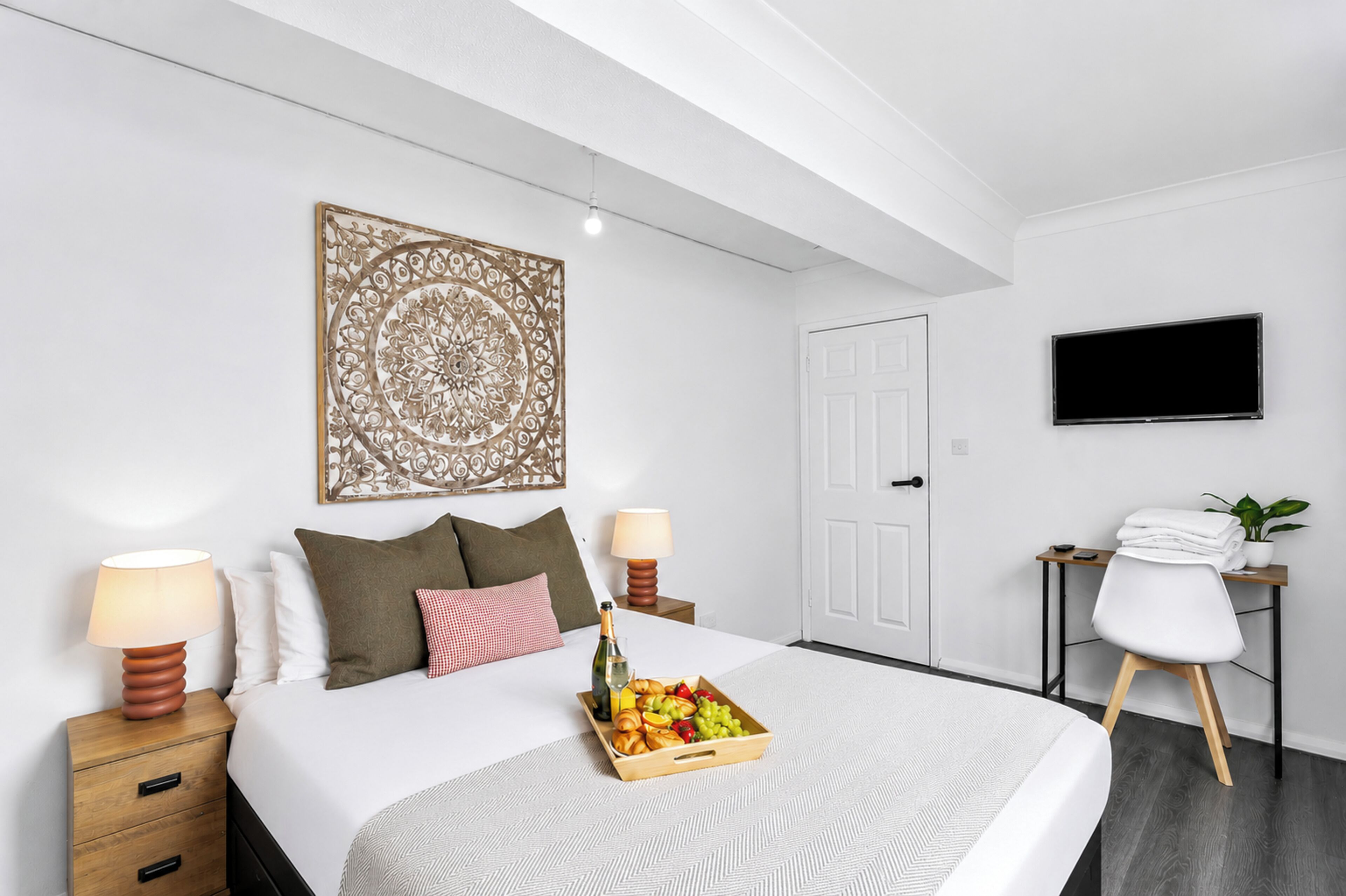 A hotel room with a bed, bedside lamps, a decorative wall piece, a TV, a desk with a chair, and a tray of fruit and wine.
