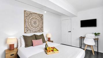 A hotel room with a bed, bedside lamps, a decorative wall piece, a TV, a desk with a chair, and a tray of fruit and wine.