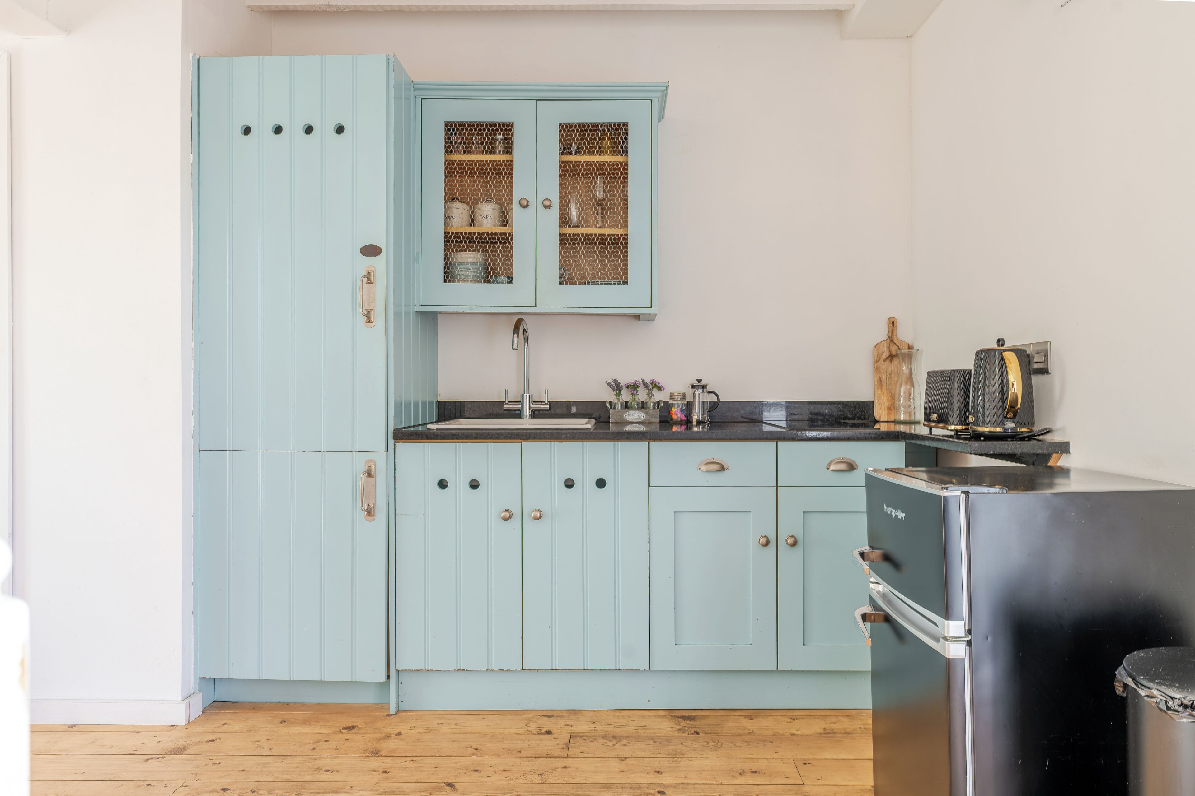 Una cocina con armarios azul claro, un refrigerador negro y piso de madera.