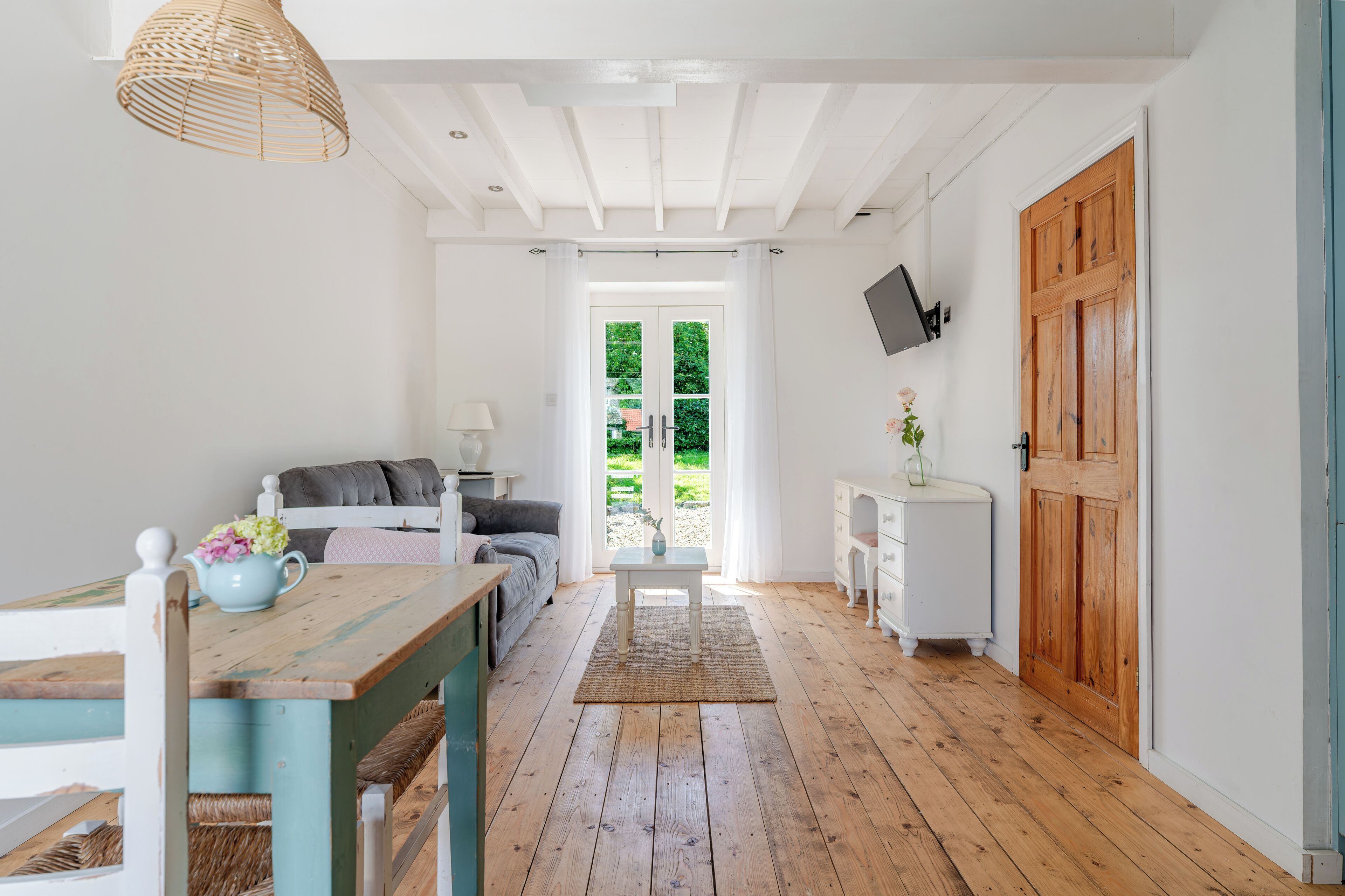 A room with a wooden floor, a table with a vase, a sofa, a TV mounted on the wall, and a wooden door.