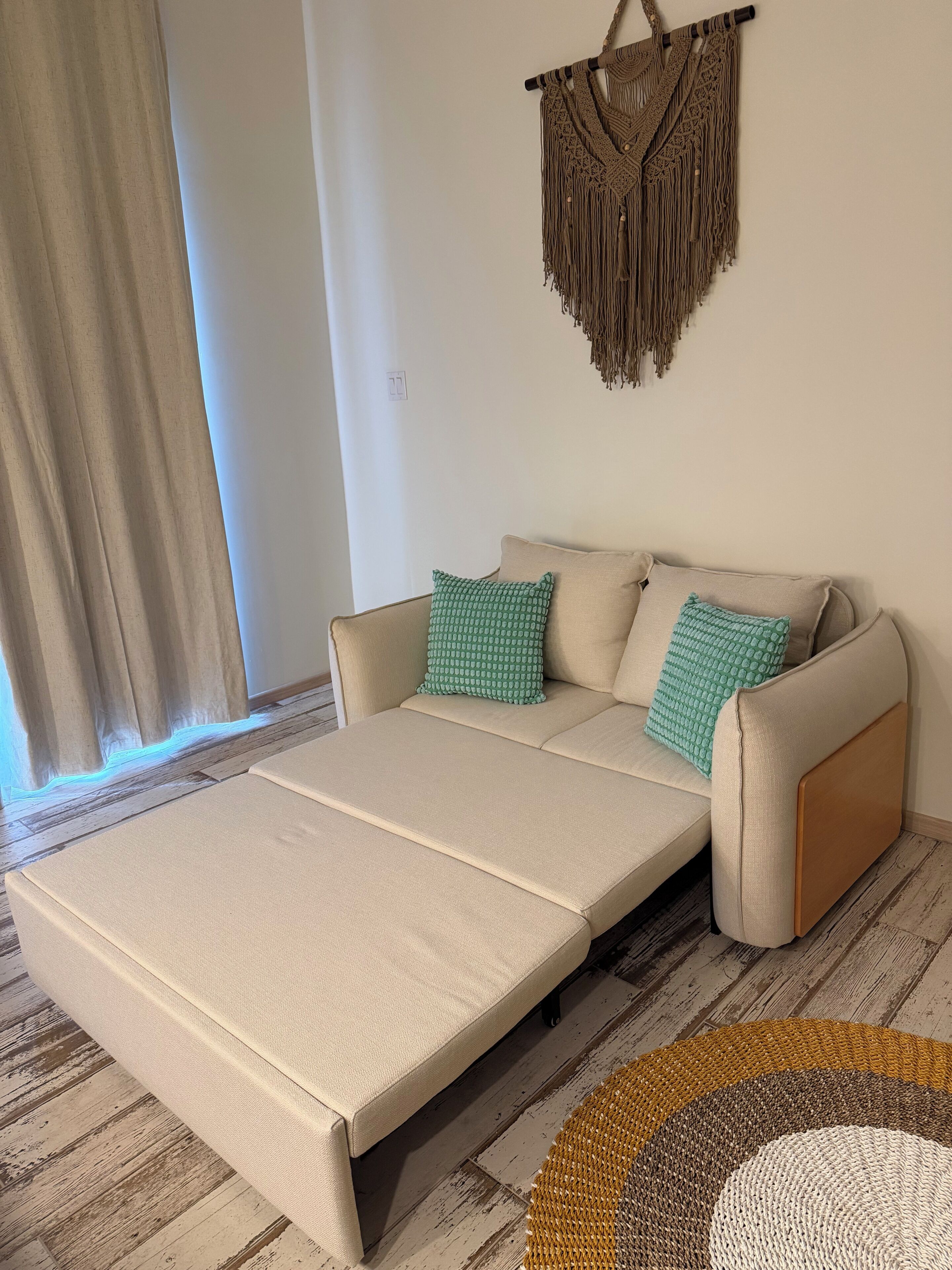 A room with a sofa bed, two pillows, a macrame wall hanging, and a patterned rug.