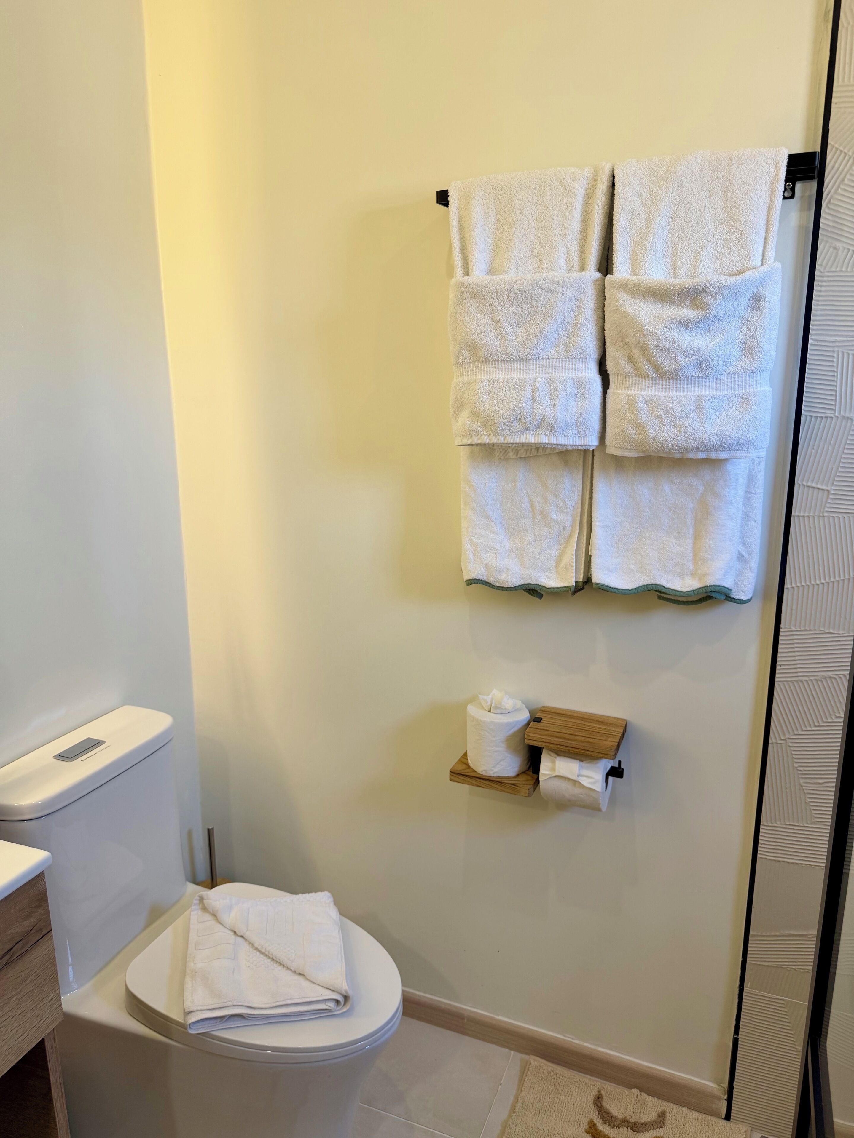 Ein Badezimmer mit Toilette, einem Handtuchhalter mit mehreren Handtüchern, einem Toilettenpapierrollenhalter und einer kleinen Holzablage.
