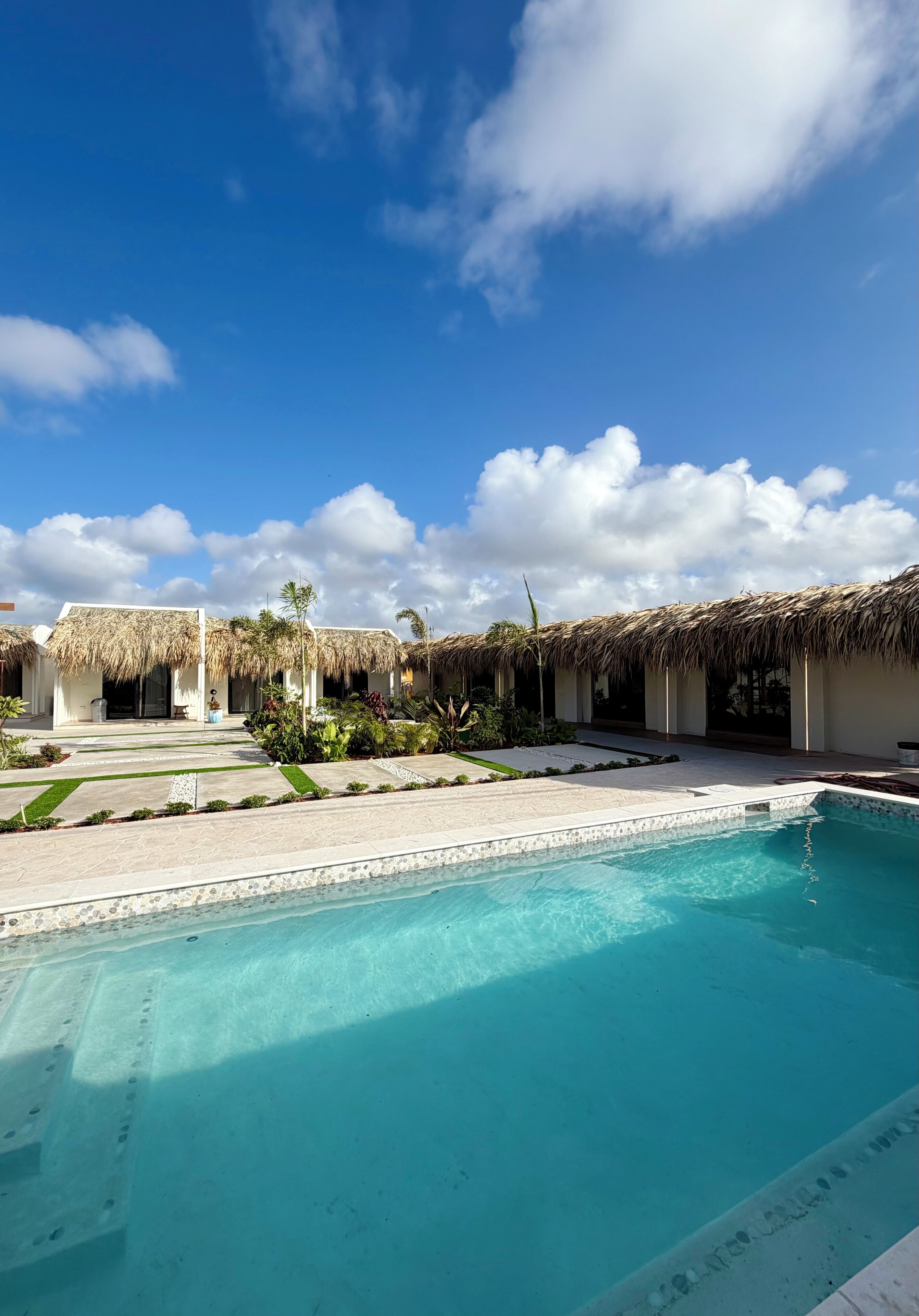 A swimming pool with a clear blue water surface, surrounded by a paved area with a thatched-roof structure.