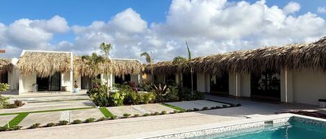 A swimming pool with a clear blue water surface, surrounded by a paved area with a thatched-roof structure.