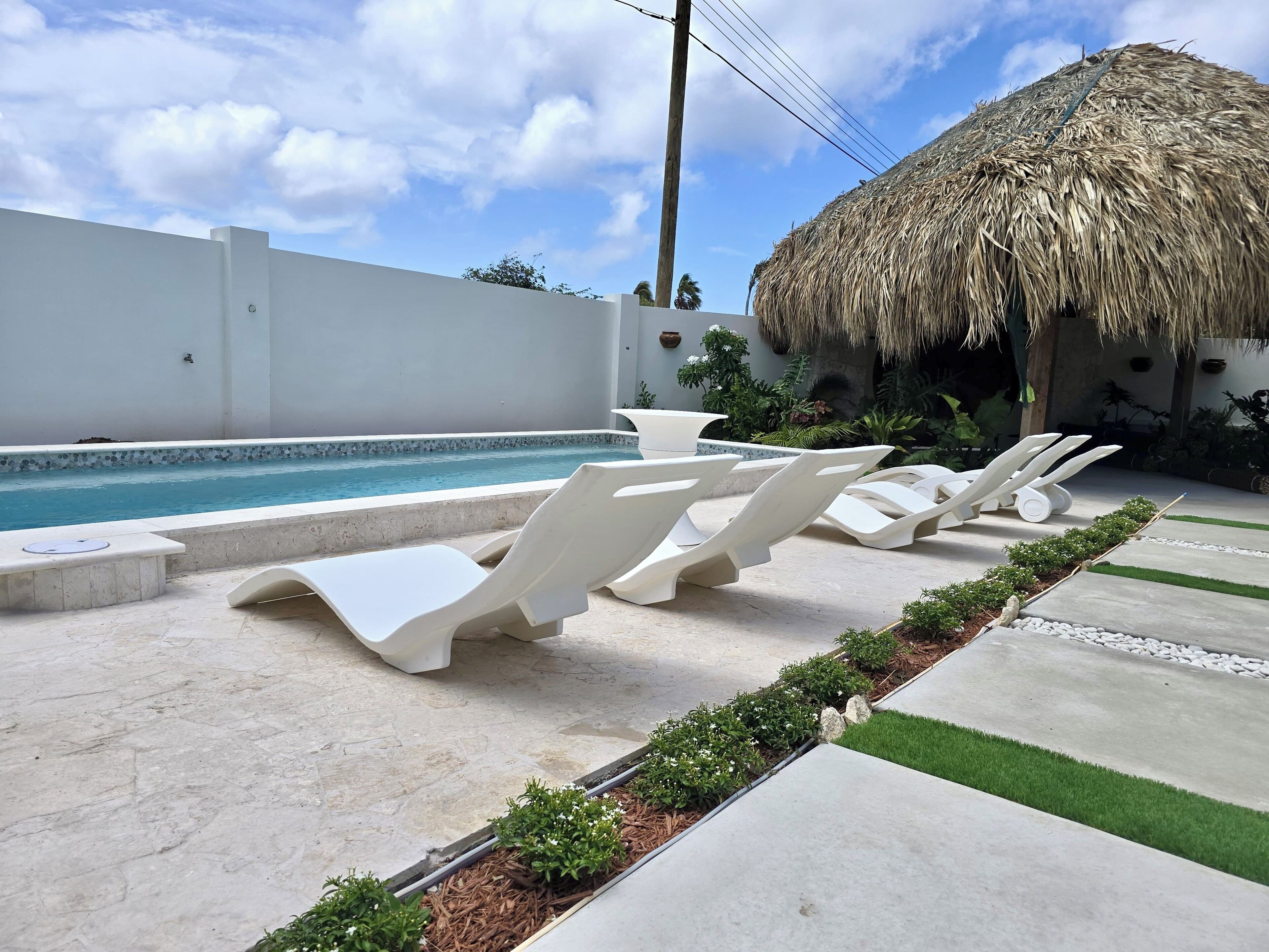 A poolside area with lounge chairs, a thatched-roof structure, and a white wall.