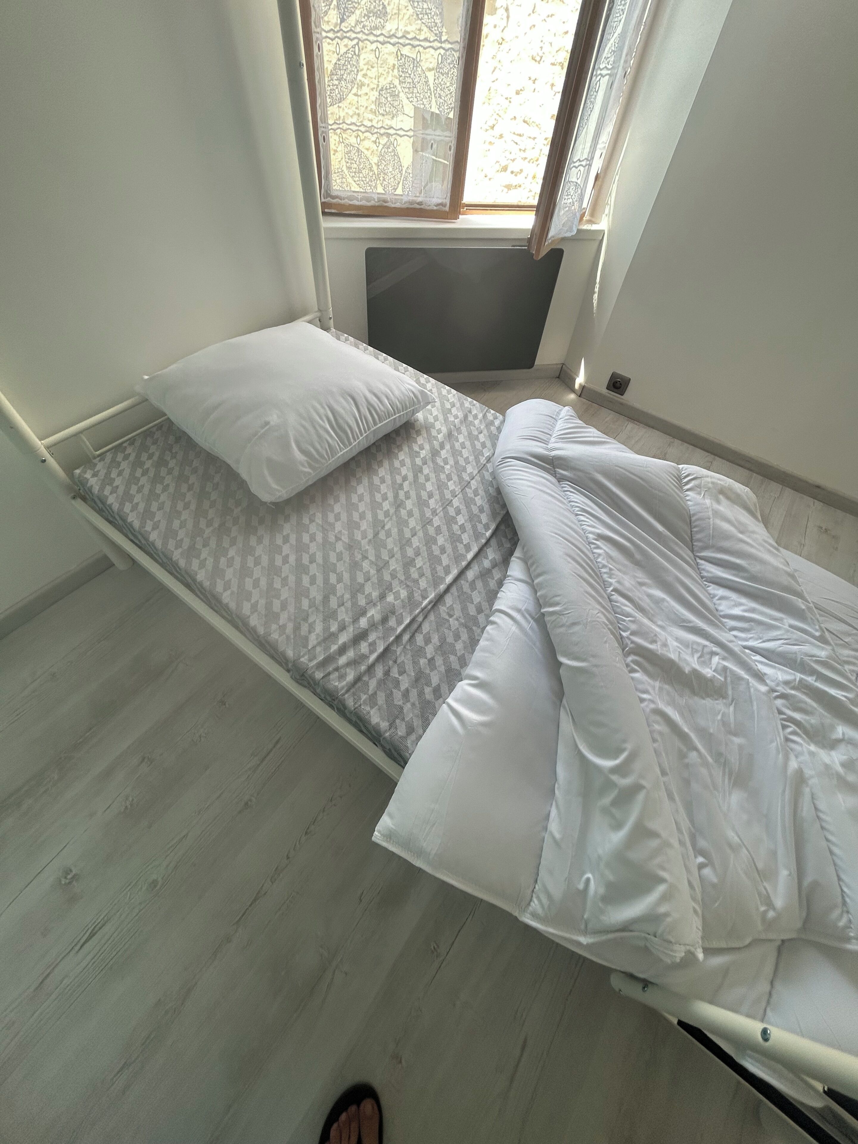 A single bed with a patterned mattress and white pillows, a window with lace curtains, and a wooden floor.