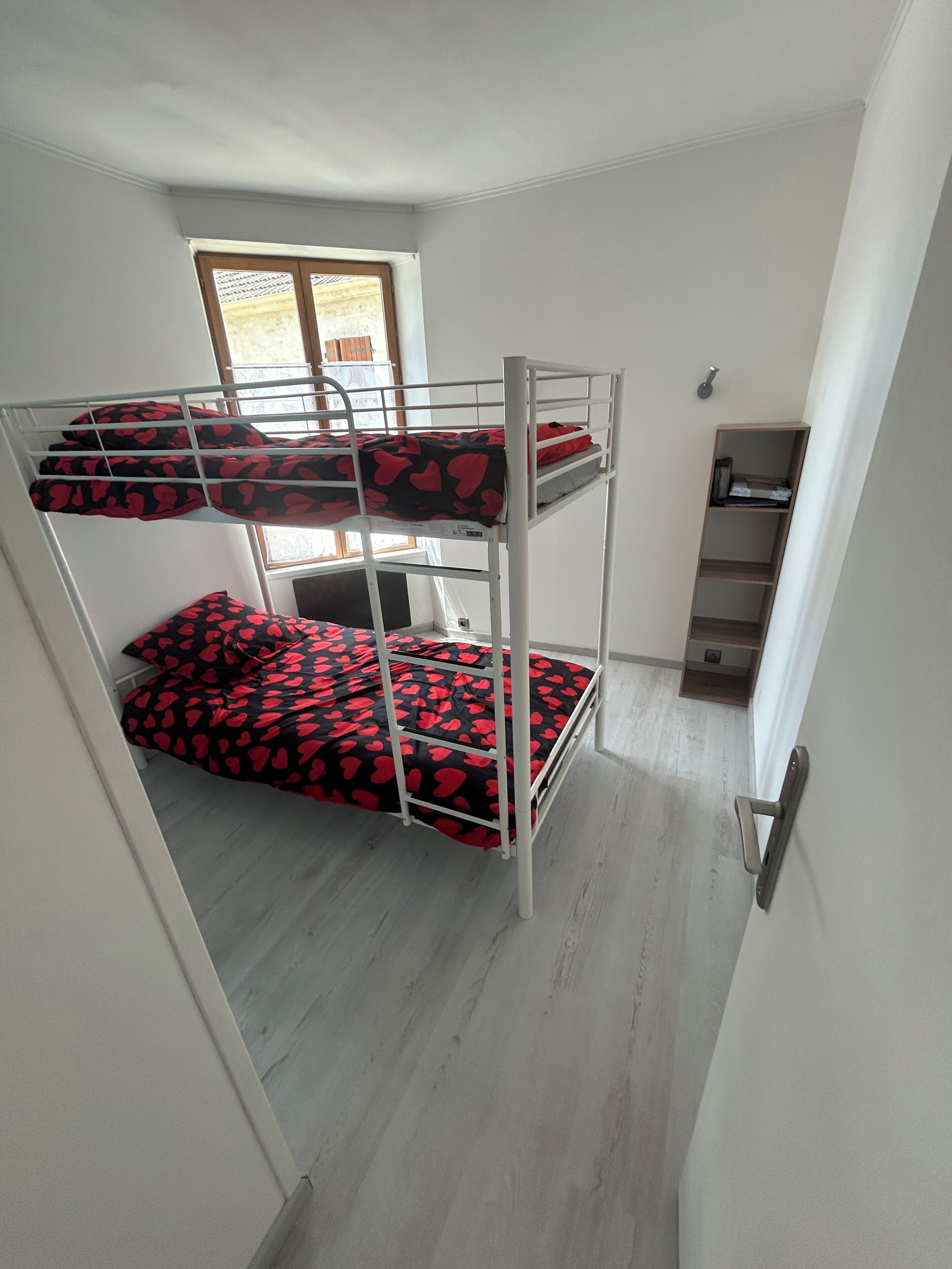 Un lit superposé avec une couvre-lit à motifs rouges et noirs, une étagère en bois et une fenêtre avec des stores.