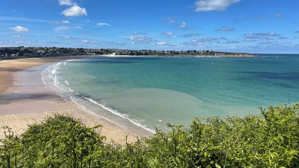 Paisagem costeira com praia, águas claras e uma linha de costa distante com edifícios.