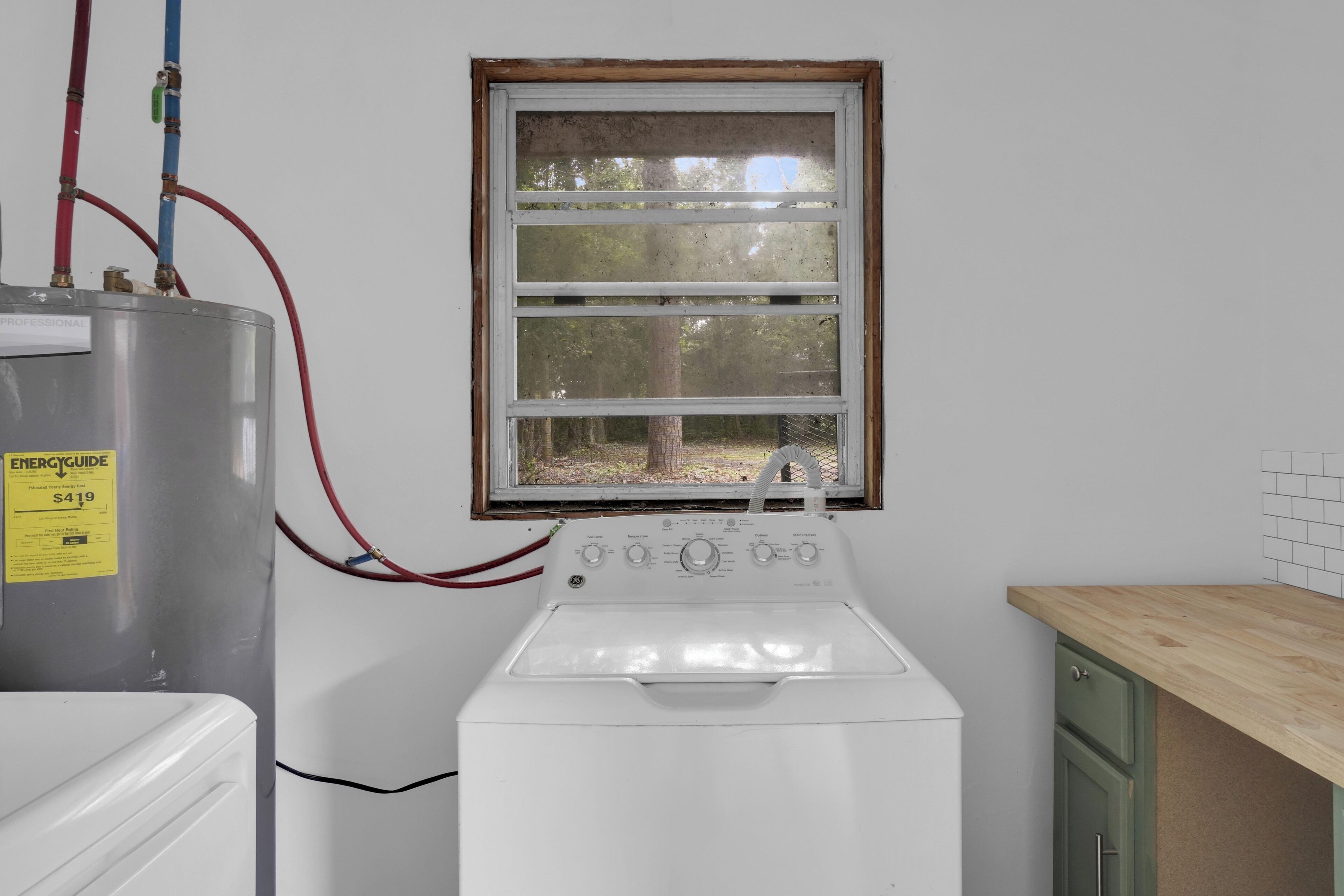 A laundry room with a water heater, a washing machine, and a window with a view of trees.