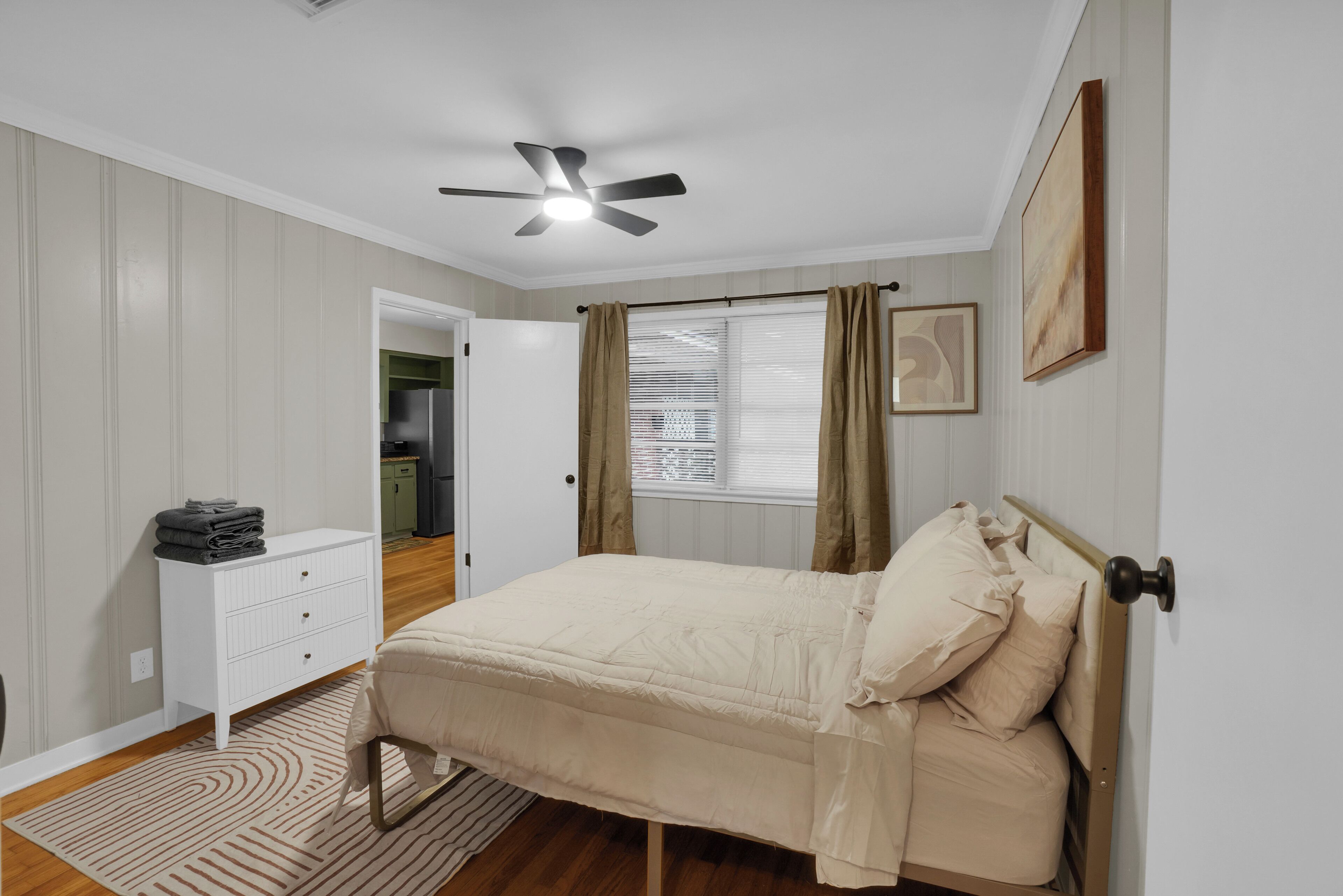 A bedroom with a bed, a ceiling fan, a window with curtains, a white dresser, and a door leading to another room.