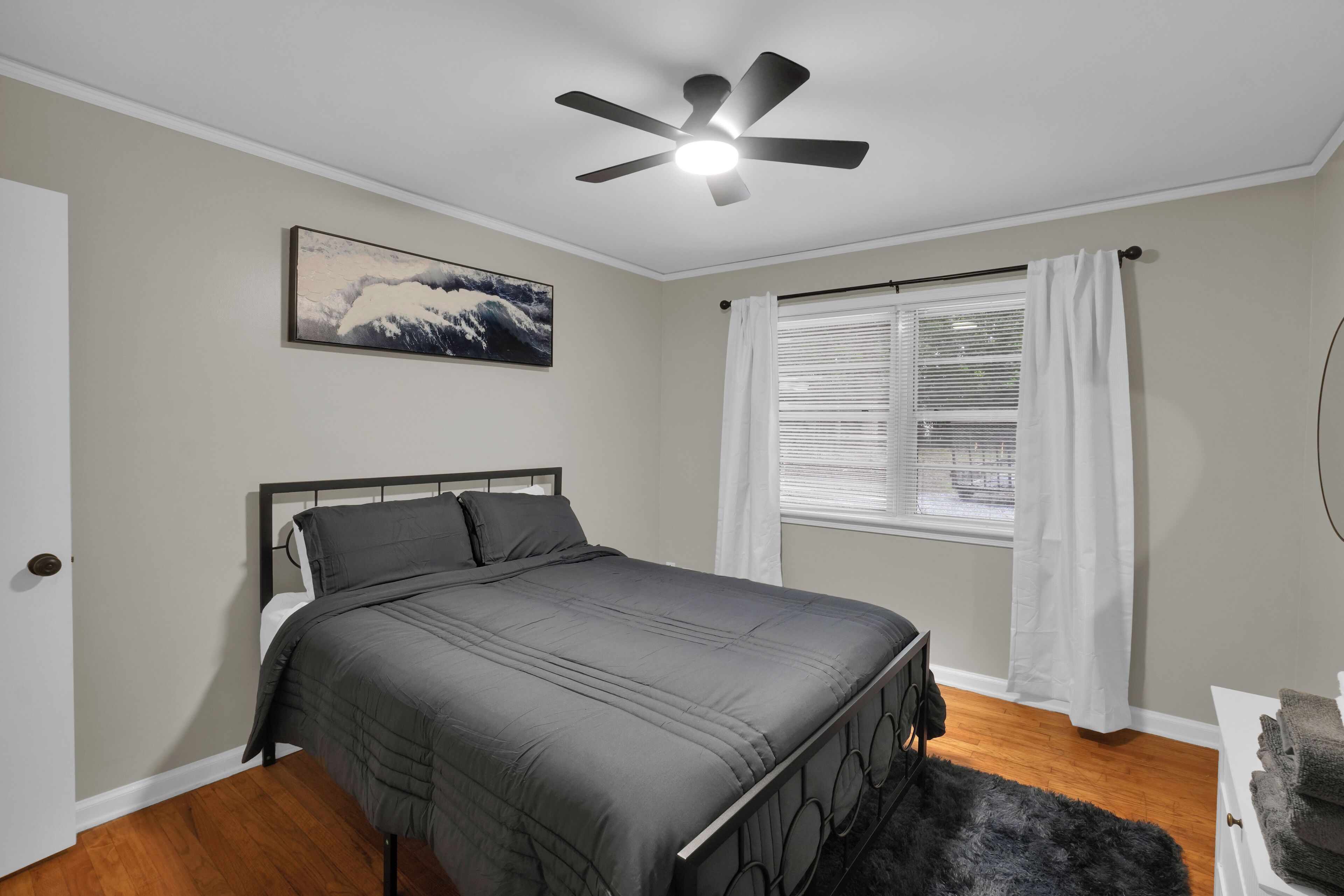 A bedroom with a bed, a ceiling fan, a window with curtains, and a painting on the wall.