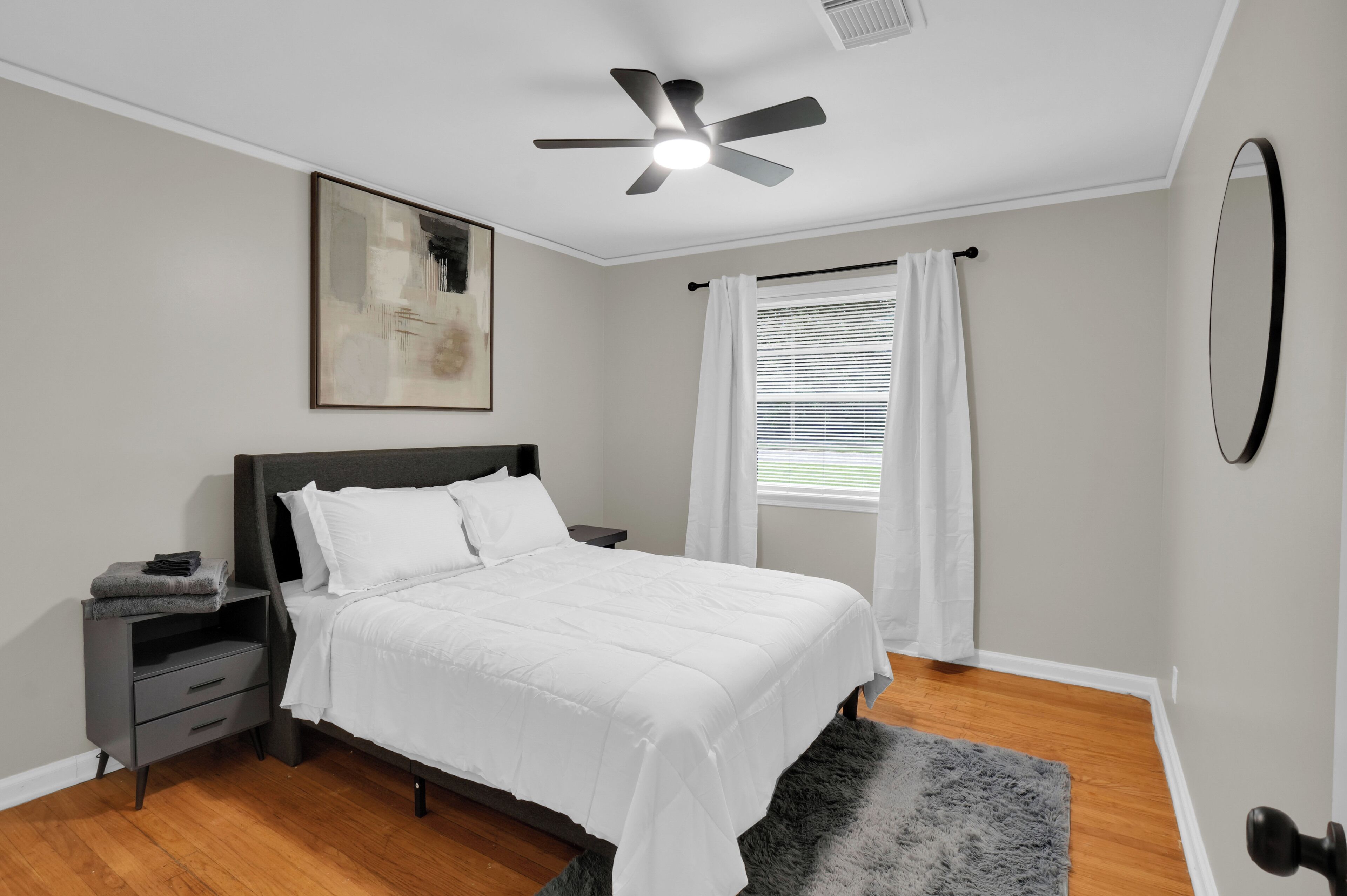A bedroom with a bed, a nightstand, a ceiling fan, a window with curtains, and a painting on the wall.