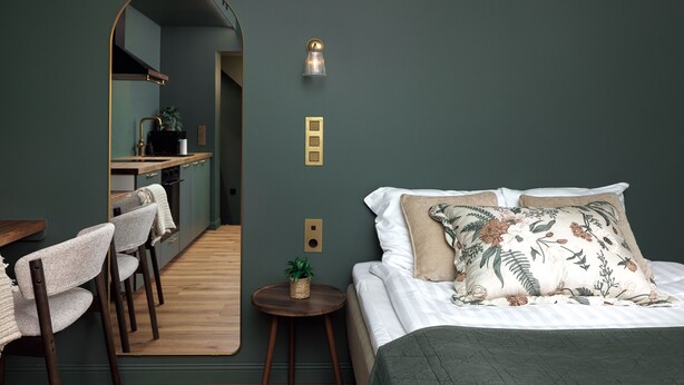 A bedroom with a bed, a mirror, a small table with a plant, and a view into a kitchen area.
