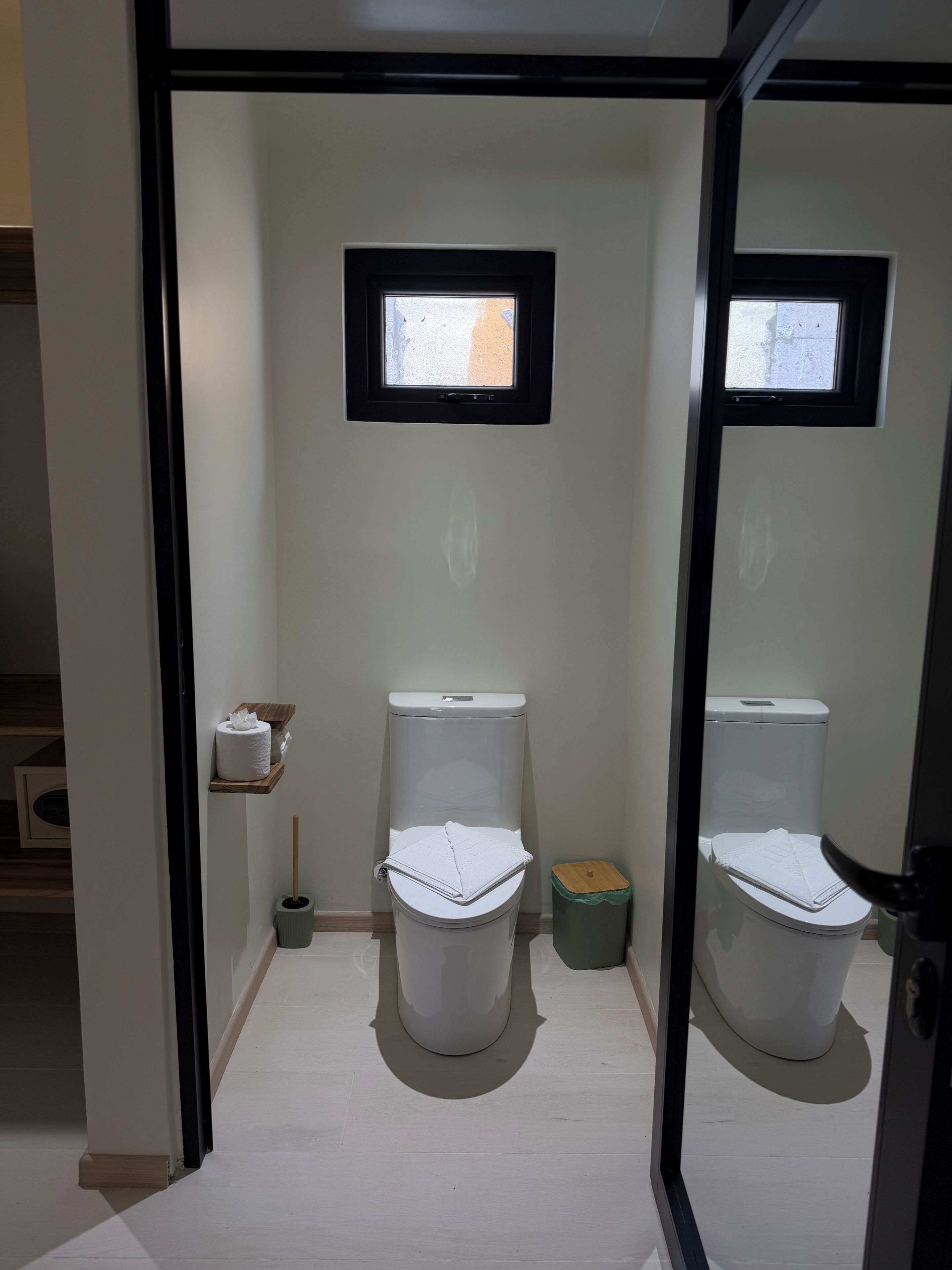 Casa de banho pequena e privada, com duas sanitas, uma janela, rolo de papel higiénico e um lixo.