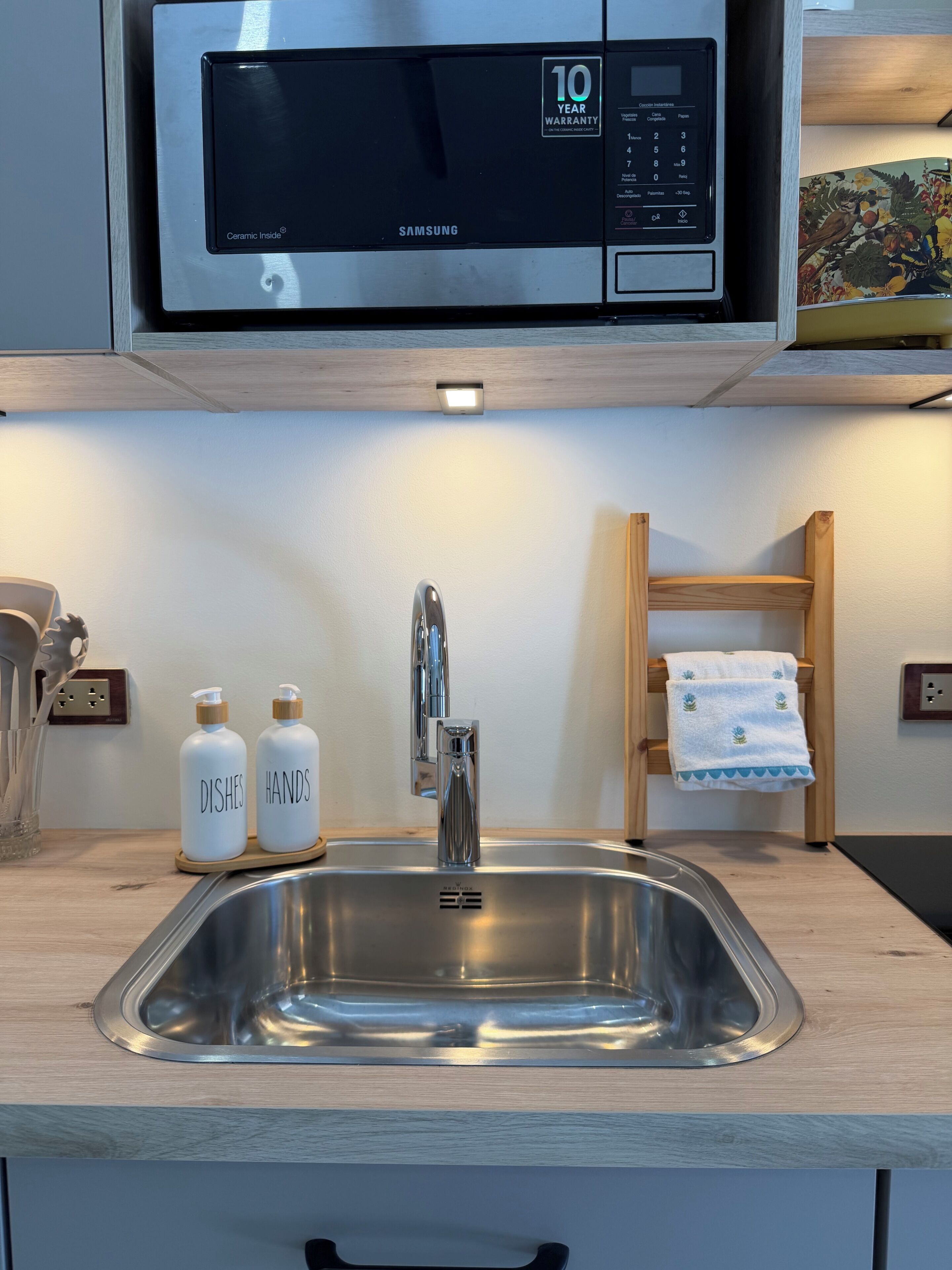 Una cocina moderna con un lavamanos de acero inoxidable, un microondas encima y dos botellas en la encimera, con las etiquetas 'DISHES' y 'HANDS'.