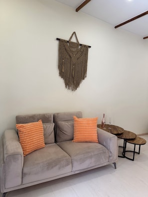 Un salon avec un canapé gris, deux coussins orange, une table basse en bois et une décoration murale en macramé.