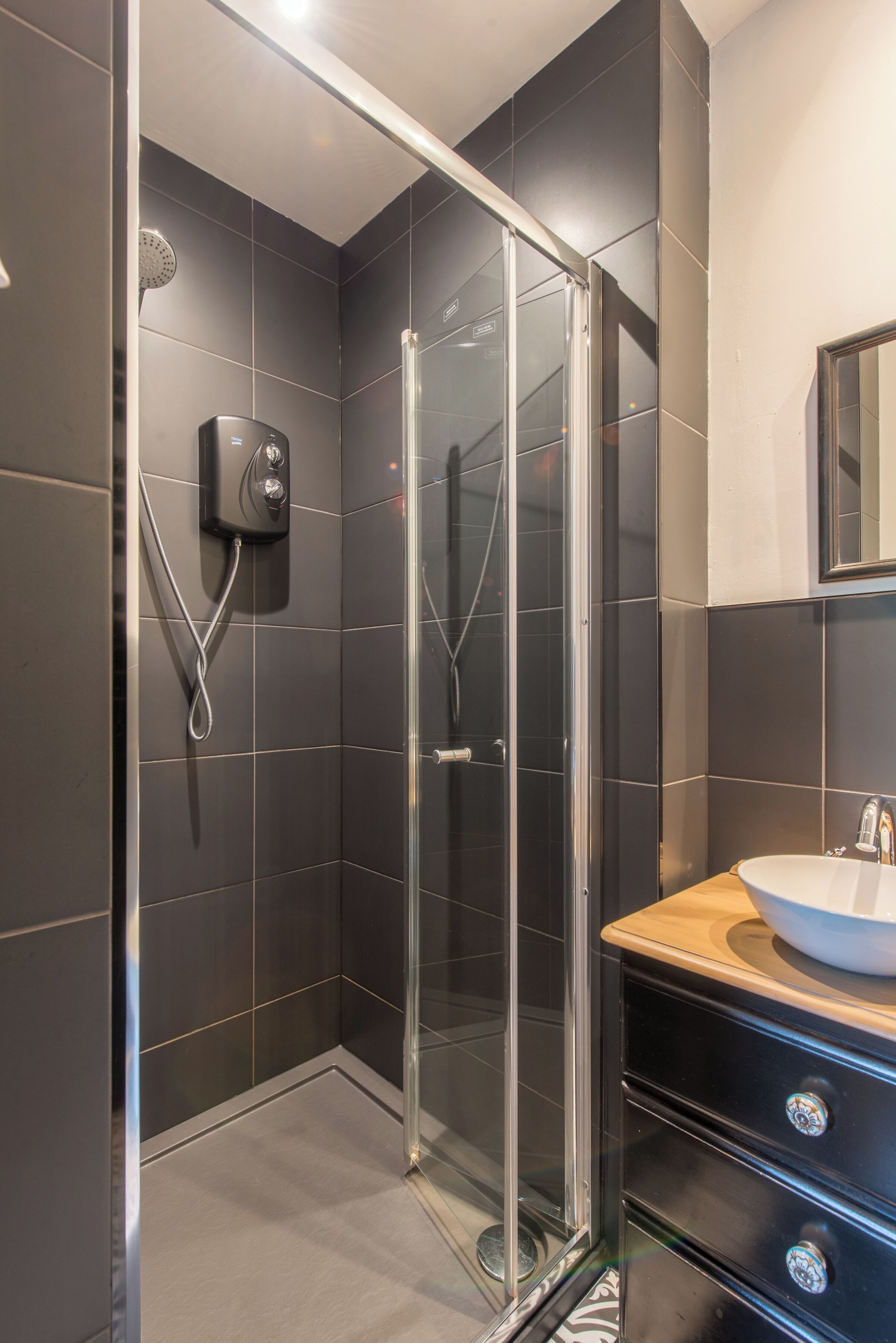 Un baño moderno con ducha envuelta en vidrio, un lavamanos con espejo y paredes de azulejos oscuros.