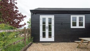 Un petit bâtiment en bois sombre, avec une porte et une fenêtre blanches, entouré de gravier et de verdure.