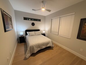 Un dormitorio con cama, mesitas de noche, ventilador de techo, ventana con persianas y estante de pared con adornos.