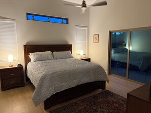 Ein Schlafzimmer mit Bett, Nachttischen, einem Fenster mit Jalousien, einem Deckenventilator und Blick durch eine Glastür.