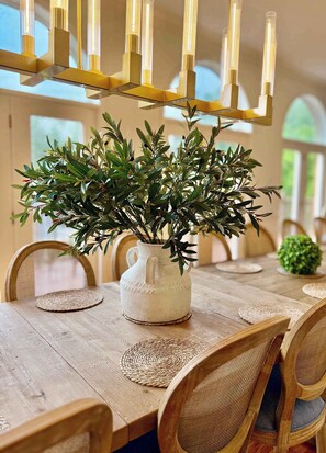 Una mesa de comedor con un mantel tejido, una jarra de cerámica blanca con follaje verde y una lámpara de araña moderna con múltiples luces.
