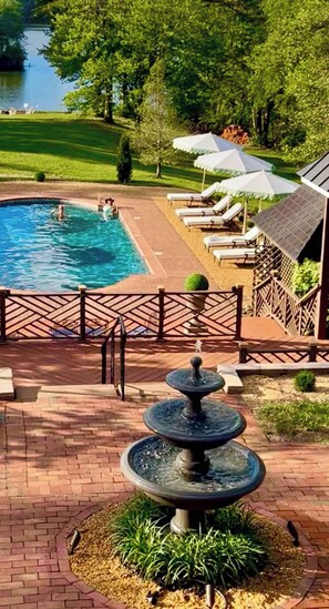 Piscina con sillas de descanso y sombrillas, rodeada por un patio de ladrillo y una fuente.