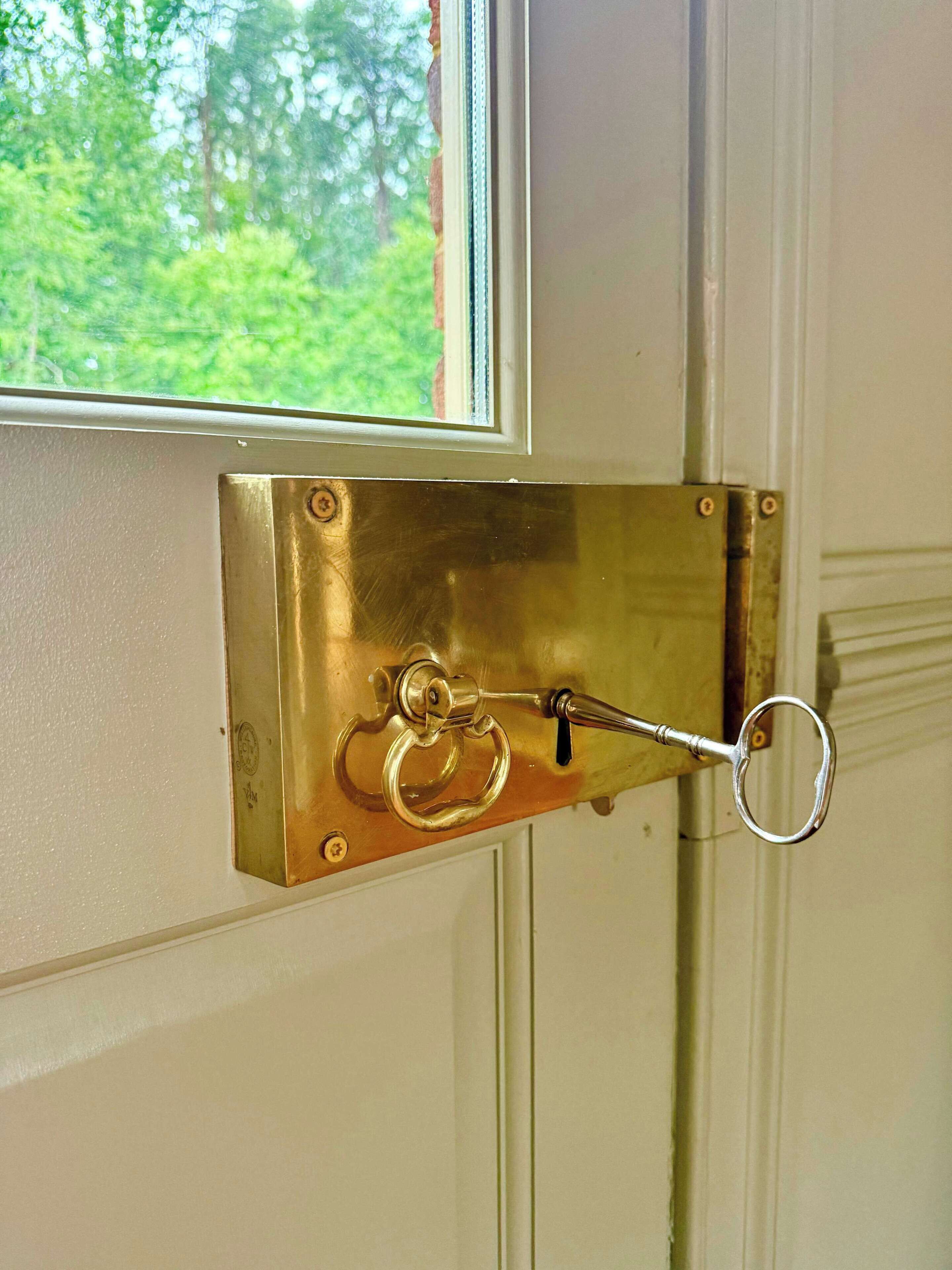 Une poignée de porte en laiton, dotée d’un trou de serrure et d’un anneau, fixée à une porte blanche surmontée d’une fenêtre.