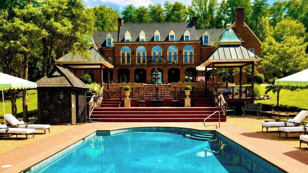 A swimming pool with a brick border, lounge chairs, and a building with arched windows and a gazebo in the background. - Private 16-Acre Lakefront Manor | Pool | Sleeps 14 (Culpeper)