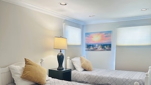 Een hotelkamer met twee bedden, een nachtkastje, een lamp, een schilderij en een raam met jaloezieën.