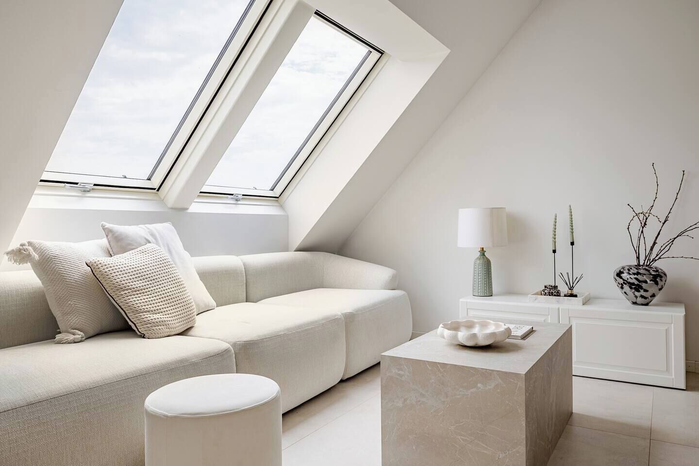 A modern loft bedroom with a sloped ceiling, a white sofa, a marble coffee table, and a nightstand with a lamp and decorative items.