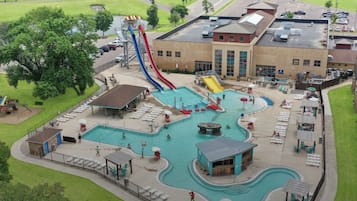 Une grande piscine extérieure dotée de plusieurs toboggans et d’un espace piscine séparé, le tout entouré d’un bâtiment à toit plat et d’un parking.