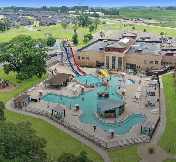 A large outdoor swimming pool with multiple slides and a separate pool area, surrounded by a building with a flat roof and a parking lot.
