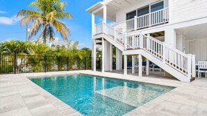 Ein Schwimmbecken mit weißer Liegefläche, ein mehrstöckiges Gebäude mit Balkon und Palmen im Hintergrund.