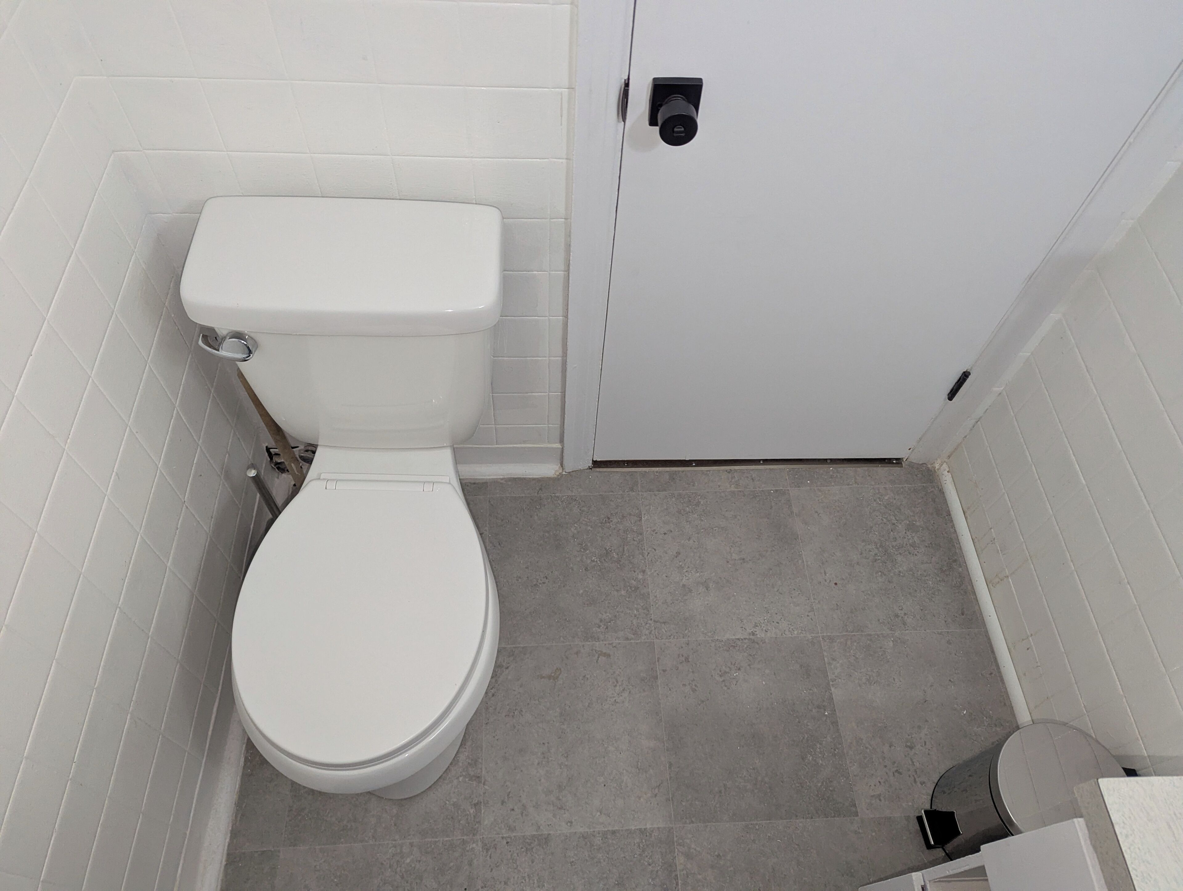 De petites toilettes blanches, avec un abattant fermé, une poignée de chasse d’eau et une porte dotée d’une serrure. Le sol est recouvert de carreaux de couleur gris clair.