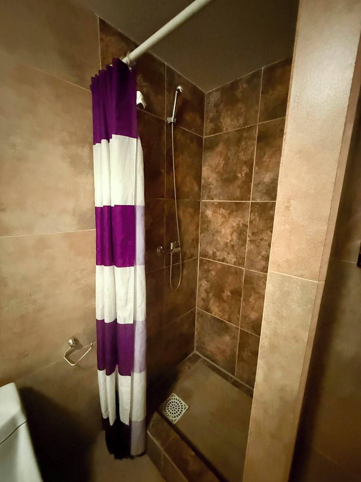 Une douche avec un rideau violet et blanc, des murs carrelés et un drain visible.