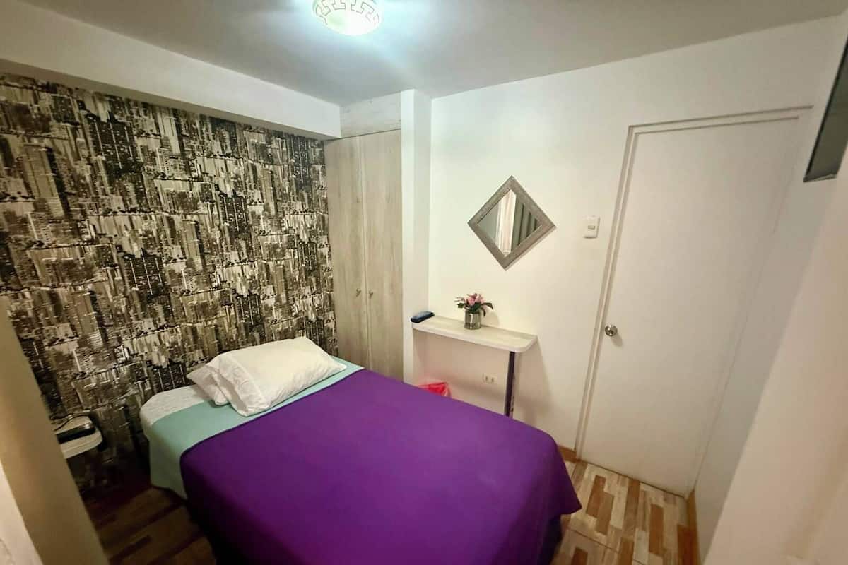 A small bedroom with a bed, a mirror, a shelf with a vase, and a patterned wall.